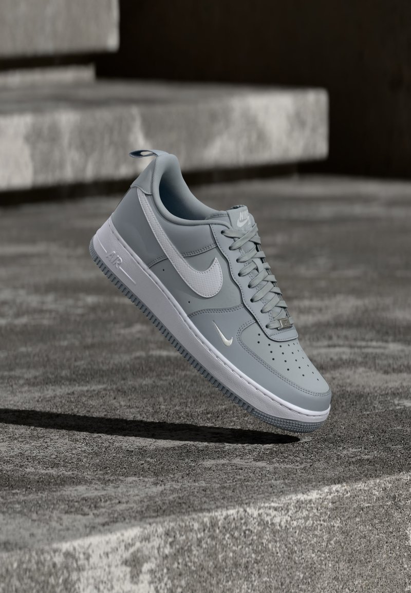 Šedý teniska Nike Air Force 1 s bielym logom swoosh, vznášajúci sa nad textúrovaným betónovým povrchom a schodmi v pozadí.