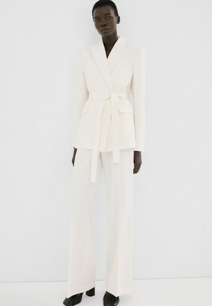 Modelo vistiendo un blazer blanco entero con cinturón y bolsillos con solapa, junto con pantalones anchos a juego, combinados con zapatos negros de tacón, posando frente a una pared blanca.