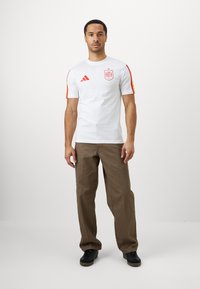 Hvit kortermet t-skjorte med røde og gule striper på ermene, med en logo og emblem på brystet. Kombinert med brune bukser og sorte sko.