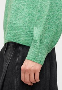 Το χέρι μιας γυναίκας ακουμπά στο πλάι, φορώντας πράσινο πλεκτό πουλόβερ και σκουρόχρωμη γκρι πλισέ φούστα, σε λευκό φόντο.