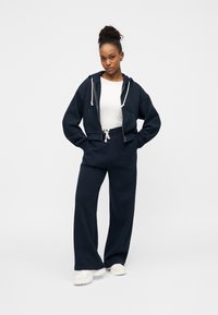 Μπλε ναυτικό cropped φούτερ με κορδόνια, συνδυασμένο με αντίστοιχο παντελόνι φαρδύ και άνετο. Και τα δύο ρούχα έχουν μαλακή υφή και άνετη εφαρμογή.