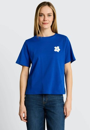 Kobieta ubrana w niebieski t-shirt z krótkim rękawem z małym białym kwiatowym wzorem na lewej piersi, w połączeniu z ciemnoniebieskimi dżinsami.