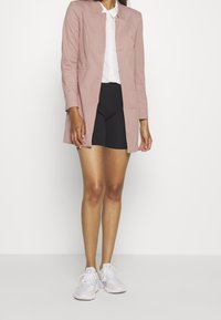 Blazer su misura rosa chiaro con maniche lunghe, apertura frontale e tasche laterali, abbinato a una camicia bianca e pantaloni corti neri, indossato con sneakers bianche.