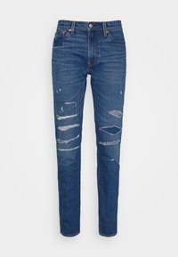 Jean en denim bleu foncé avec des détails déchirés, présentant des zones légèrement décolorées et une coupe droite. Style à cinq poches avec quincaillerie en métal.