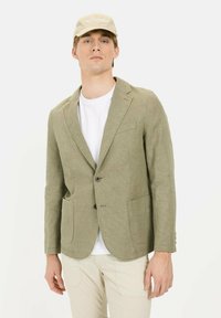 Lichtgroene linnen blazer met twee voorkruiszakken, sluiting met één knoop en notch lapel, gedragen over een wit T-shirt en aangevuld met een beige pet.