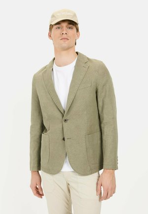 Lichtgroene linnen blazer met twee voorkruiszakken, sluiting met één knoop en notch lapel, gedragen over een wit T-shirt en aangevuld met een beige pet.