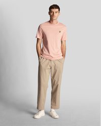 T-shirt en coton rose clair à manches courtes, avec un petit logo jaune. Assorti avec un pantalon chinos beige et des baskets blanches. Design simple et décontracté.
