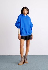 Blue oversized hoodie with a front pocket, paired with a black pleated skirt and tan ankle boots with laces. Smooth fabric textures.