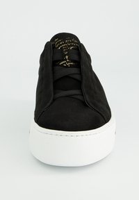 Schwarze Wildledersneaker mit dicker weißer Sohle, versteckten Schnürsenkeln und goldener, geschwungener Schrift auf der Zunge, von vorne betrachtet.