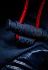 Musta ja punainen säädettävä narulukko, jossa on painatukset "JK1T TM" ja "JACK1T TM", kiinnitettynä sileälle laivastonsiniselle kangaspohjalle.