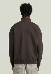 Donkerbruine sweater met een hoge kraag, afhangende schouders, ribgebreide mouwen en onderrand. Bevat een vierkant label op de achterkant.