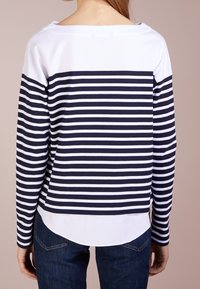 Persona che indossa una maglietta a righe orizzontali blu navy e bianca a maniche lunghe con orlo bianco a strati, abbinata a jeans blu scuro, vista da dietro.