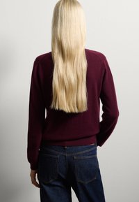 Maglione bordeaux con una vestibilità aderente e polsini a coste, abbinato a jeans blu scuro con tasche posteriori e cuciture gialle. Lunghi capelli biondi scorrono lisci.