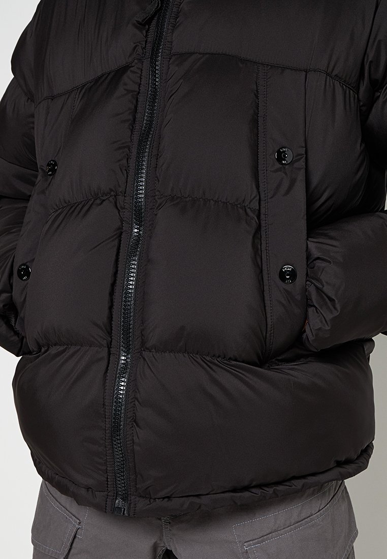 Schwarze Steppjacke mit Reißverschluss vorne, zwei großen Knöpfen und einem gesteppten Design. Das Material ist glatt und hat eine weiche Textur.