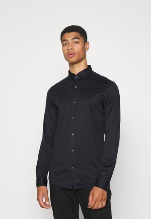 Chemise noire à manches longues avec un col boutonné, fabriquée en tissu lisse, présentant une coupe ajustée et des accents de boutons sur le devant.
