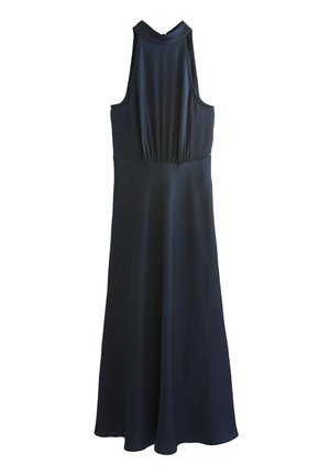 Kvinde i en sort ærmeløs halter-neck kjole og sorte hæle, poserer foran en grå baggrund, mens hun let holder kjolen.