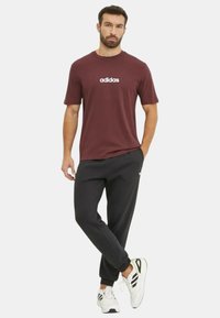 T-shirt in cotone color bordeaux con logo "adidas" bianco, abbinata a pantaloni della tuta neri e sneakers bianche con accenti e lacci neri.