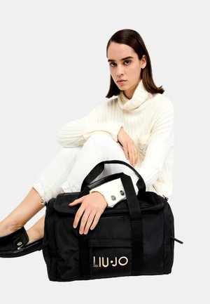 Borsa duffel nera con tessuto testurizzato, manici doppi, tasca frontale e logo dorato "LIU.JO". Modella che indossa un maglione lavorato off-white e pantaloni bianchi.