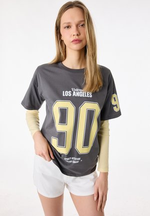 Młoda kobieta o długich blond włosach nosi szary t-shirt z napisem "Los Angeles 90" na bladoróżowym długim rękawie i białych szortach, stojąc na białym tle.