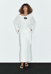 White linen dress with wide sleeves and a waist tie. Model wears black pendant necklace and brown sandals. Background is light gray.