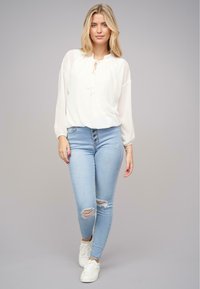 Hvid, semi-gennemsigtig bluse med bindebånd i halsen, lange ærmer, parret med lyseblå revnede skinny jeans og hvide sneakers.