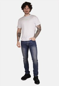 T-shirt di cotone bianco con un piccolo logo verde, abbinata a jeans aderenti blu e sneakers nere. Modello in posa neutra.