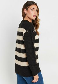 Maglione a righe nere e crema con lavorazione a maglia testurizzata, caratterizzato da bottoni tondeggianti sulle spalle. Il modello lo indossa con jeans blu.