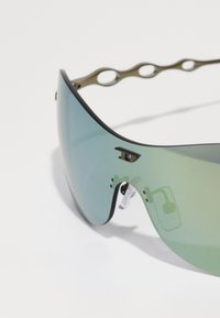 Lunettes de soleil surdimensionnées teintées de vert avec un verre lisse et courbé, une monture noire et un design de branches en métal ondulées distinctif.