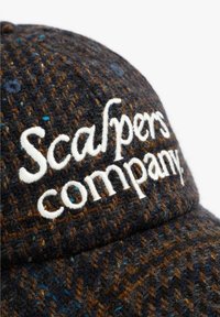 Casquette en laine avec un motif pied-de-poule bleu marine et marron. Texte brodé blanc "Scalpers company". Tissu texturé avec une visière courbée.