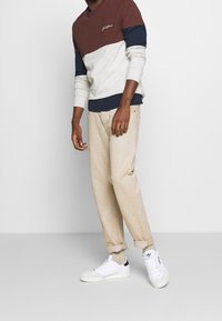 T-shirt i bruna, grå och marinblå färgblock, med lång ärm; ihop med beige byxor och vita träningsskor.