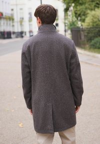 Man som bär en mörkgrå yllekappa och står på en trottoar i staden med händerna avslappnat vid sidorna, vänd bort från kameran.