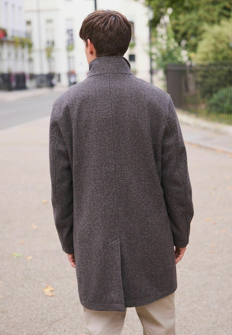 Man som bär en mörkgrå yllekappa och står på en trottoar i staden med händerna avslappnat vid sidorna, vänd bort från kameran.