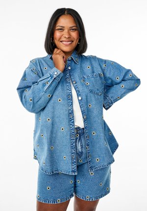 Ensemble chemise en denim et shorts avec un tissu bleu orné de embellissements circulaires. La chemise possède une poche et une fermeture à boutons.