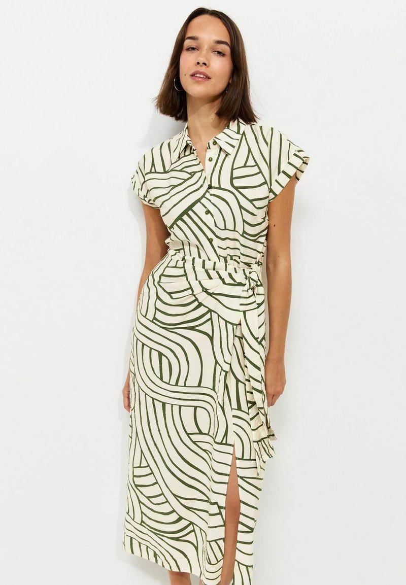 Woman wearing a white short-sleeve buttoned dress with green abstract line patterns and a waist tie standing against a white background.
