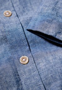 Lichtblauwe linnen shirt met knoopsluiting, met een gestructureerd oppervlak en een opgerolde mouw, die subtiele variaties in schaduw laat zien.