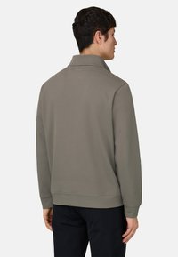 Homme vu de dos portant un pull à manches longues gris avec col et un pantalon noir, sur un fond uni.