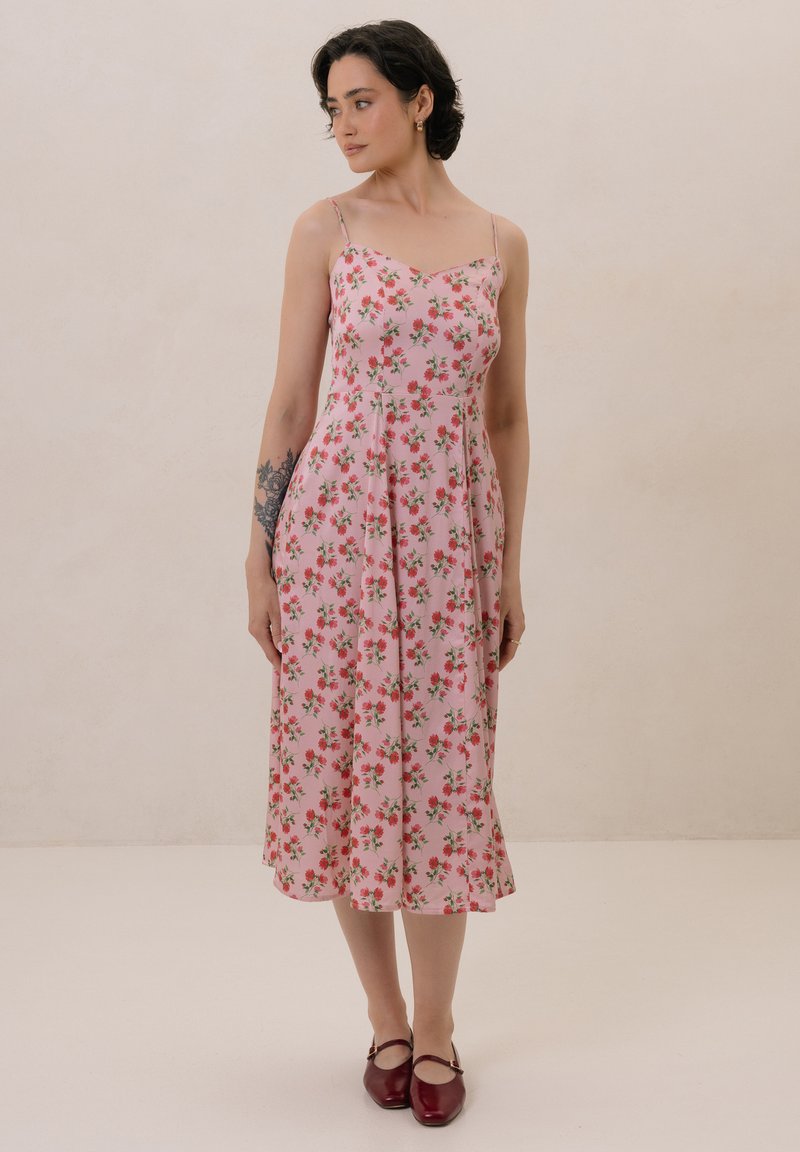 Różowa sukienka kwiatowa z cienkimi ramiączkami, w fasonie A-line, uszyta z lekkiego materiału. Charakteryzuje się małymi czerwonymi kwiatkami i zielonymi liśćmi w rozproszonym wzorze.