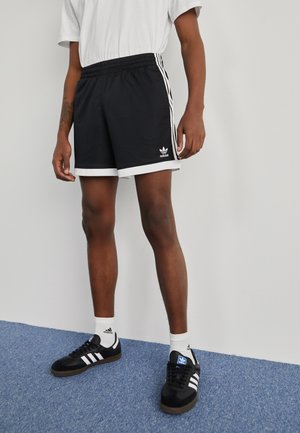 Osoba ubrana w czarne spodenki Adidas z białym obszyciem, białe skarpetki Adidas i czarne sneakersy Adidas, stojąca na niebieskim dywanie.
