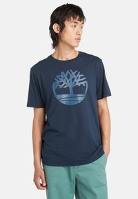 La camiseta de algodón de color azul marino cuenta con un gran logo de árbol en color azul claro en el frente, mangas cortas y un cuello redondo.