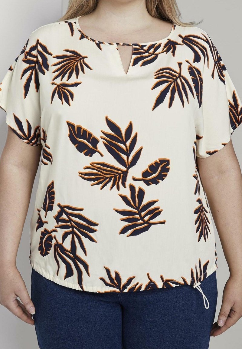 Blusa beige chiaro con stampa di foglie navy, dotata di scollatura a fessura e maniche corte. La parte inferiore ha un cordoncino regolabile.