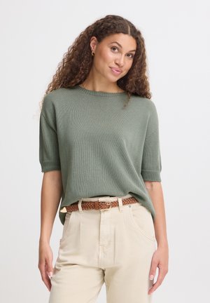 Woman with curly hair wearing a green short-sleeve sweater, beige pants, and a brown braided belt standing against a plain background.