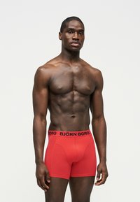 Homme musclé debout, portant un boxer rouge Björn Borg sur un fond clair uni, regardant devant avec une expression neutre.