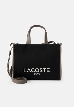 Borsa in tessuto nera con dettagli in pelle grigia, dotata di doppi manici superiori e una tracolla rimovibile. Logo "LACOSTE 1933" in bianco sulla parte anteriore.