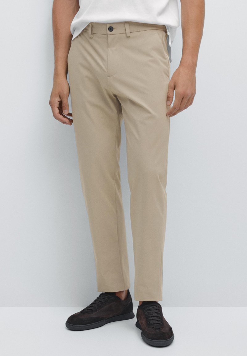Béžové kalhoty na míru s hladkou texturou, s knoflíkovým zapínáním a bočními kapsami, nošené s tmavě hnědými teniskami.