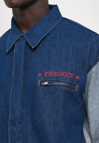Veste en denim avec des manches claires contrastantes, texte rouge "FIORUCCI" et étoiles au-dessus d'une poche zippée horizontale sur la poitrine.