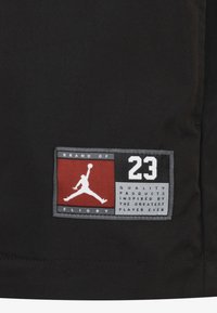Svart stoff med et rektangulært merke som har røde, hvite og grå akcenter. Merket inneholder Jumpman-logoen og tekst i en fet skrift.