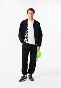 Musta kevyt takki ja juoksuhousut elastisilla hihansuilla valkoisen poolopaidan päällä, jossa on suuri "LACOSTE"-logo. Vihreä lakki käsissä.