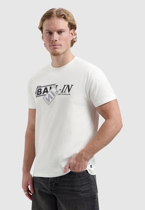 Jonge man met licht gekruld haar draagt een wit T-shirt met print en zwarte jeans, staat met één hand in zijn zak tegen een effen achtergrond.