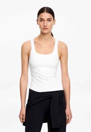 Femme aux cheveux foncés attachés, vêtue d'un débardeur blanc sans manches et d'un pantalon noir avec une veste noire nouée autour de la taille.