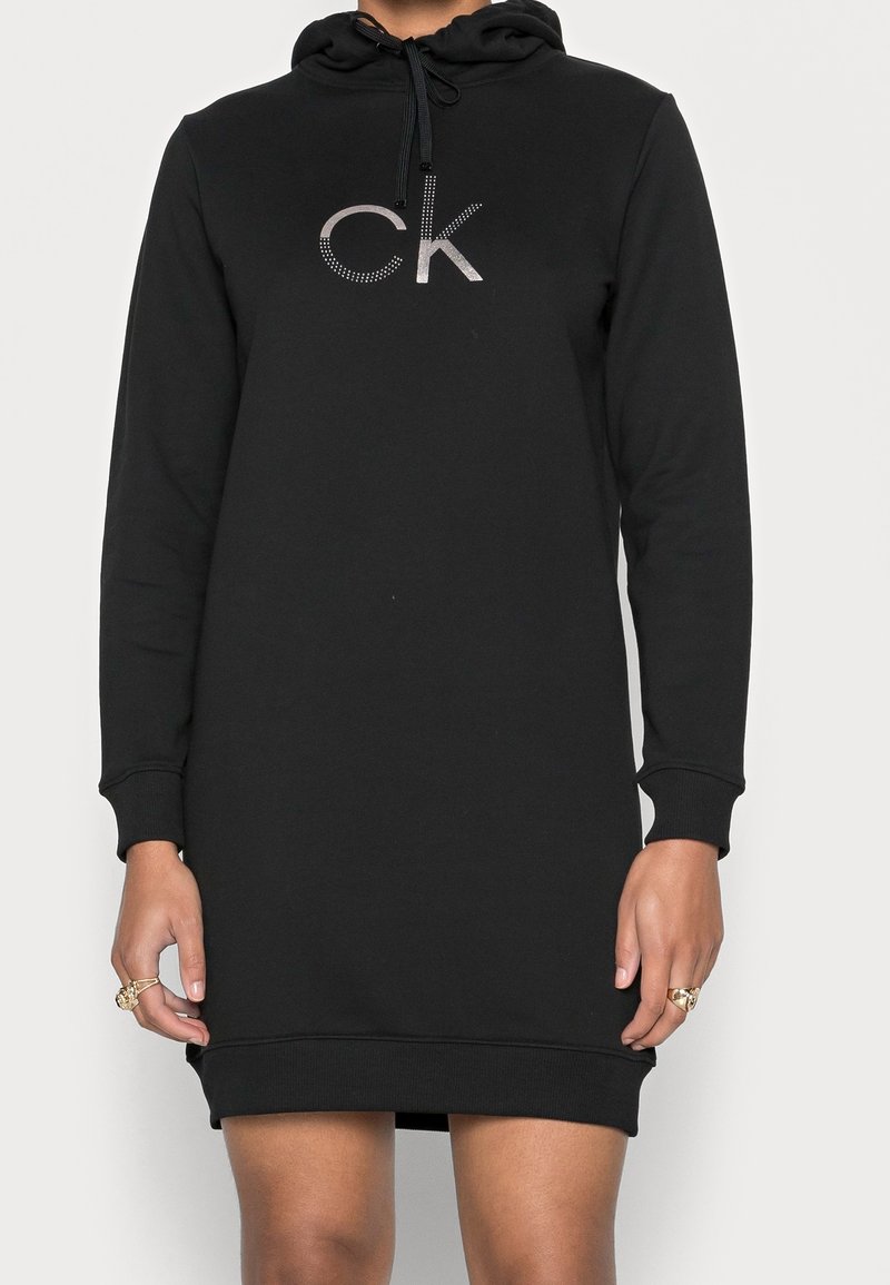 Robe à capuche noire à manches longues avec un petit logo métallique "ck" sur la poitrine, portée par une personne montrant des mains avec des bagues.